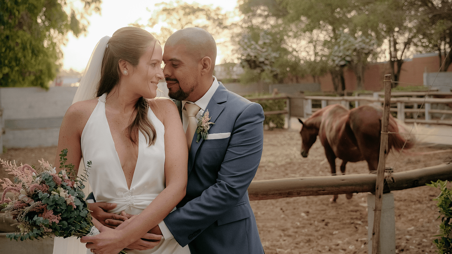 Una novia y un novio abrazados al atardecer en un rancho, con un caballo detrás